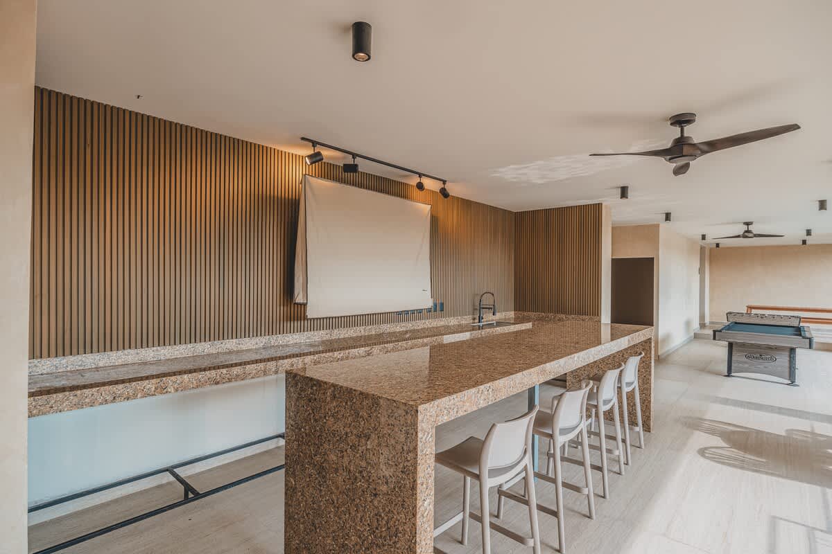 A spacious outdoor bar area features a long, speckled granite counter with white bar stools, backed by a slatted wooden wall with a projector screen. Recessed lighting and a ceiling fan illuminate the space, with game tables visible in the distance.