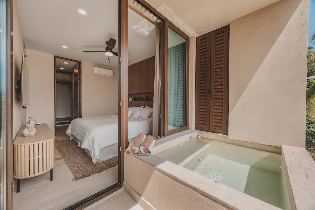 Bedroom with a white-linened bed and a wooden dresser, featuring a sliding glass door that opens to a balcony with a built-in hot tub and a view of the surrounding landscape.