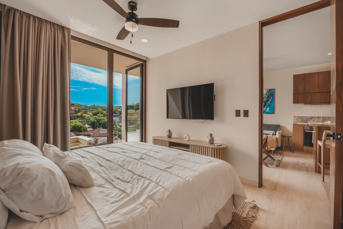 A bright bedroom with a white-linened bed facing a wall-mounted TV above a light wooden console, featuring large glass doors that open to a balcony with lush green views. A dark ceiling fan hangs overhead.