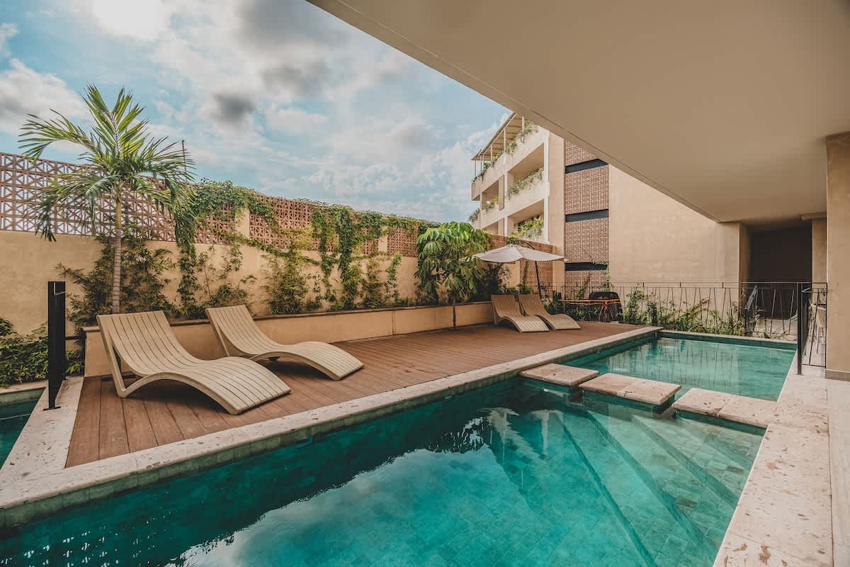 Inviting swimming pool with clear turquoise water, featuring a wooden deck with two sleek beige lounge chairs and a white umbrella. The area is framed by a stone border and a wall covered in climbing vines, with palm trees adding to the tropical ambiance.