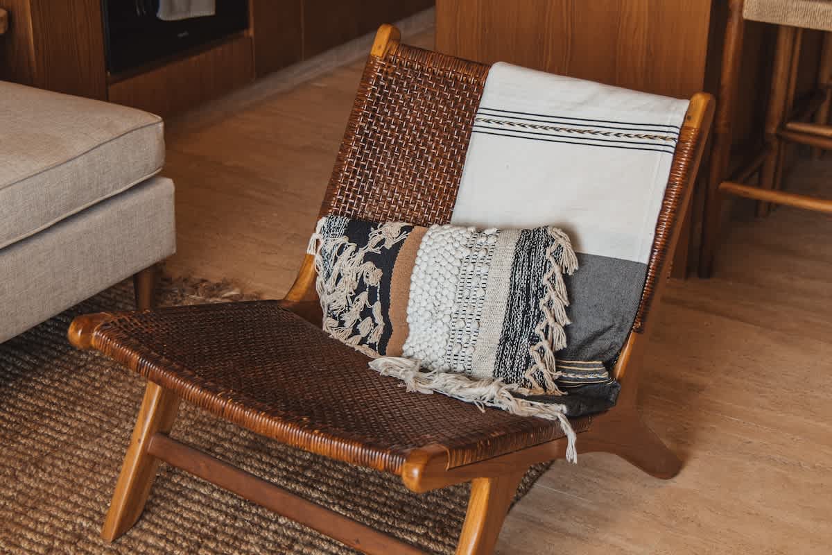 A full view of a wooden lounge chair with woven cane seat and back, featuring a cream and black striped throw and a textured decorative pillow with tassels, set on a natural fiber rug.