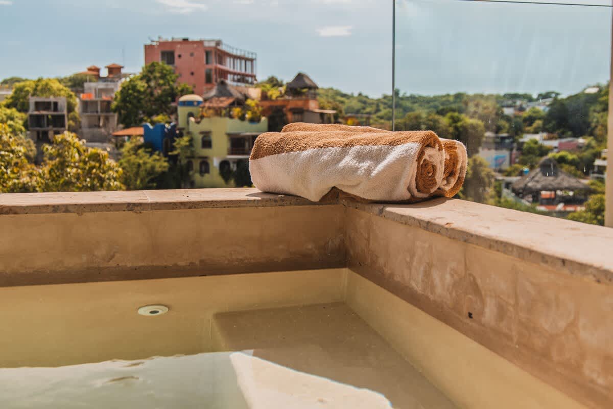 An outdoor hot tub on a balcony with a clear glass railing, featuring two neatly rolled brown and white striped towels on the edge, overlooking a scenic town with colorful buildings and lush trees.