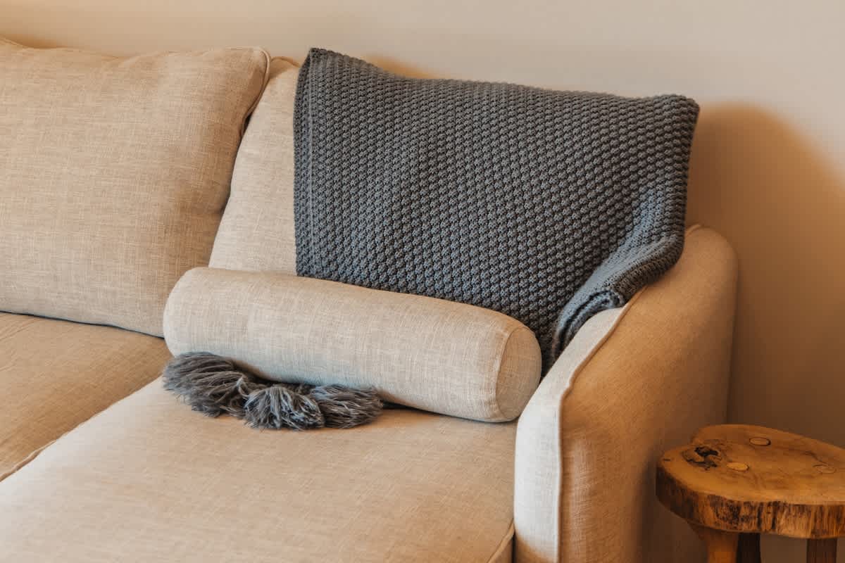 A comfortable beige sofa features a grey knitted throw pillow and a matching bolster pillow with decorative tassels, suggesting a cozy seating area.