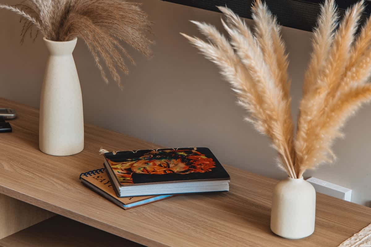 Two white ceramic vases filled with dried pampas grass flank a stack of decorative books on a light wood console table, creating a warm and natural decor accent.