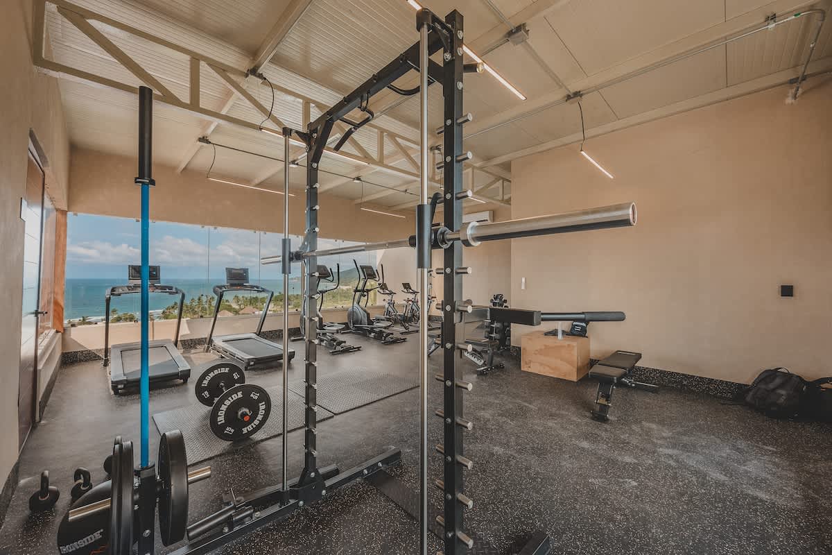 A well-equipped gym offers both strength training with a power rack and free weights, and cardio machines, all set against a backdrop of a vibrant ocean and town vista visible through large windows.