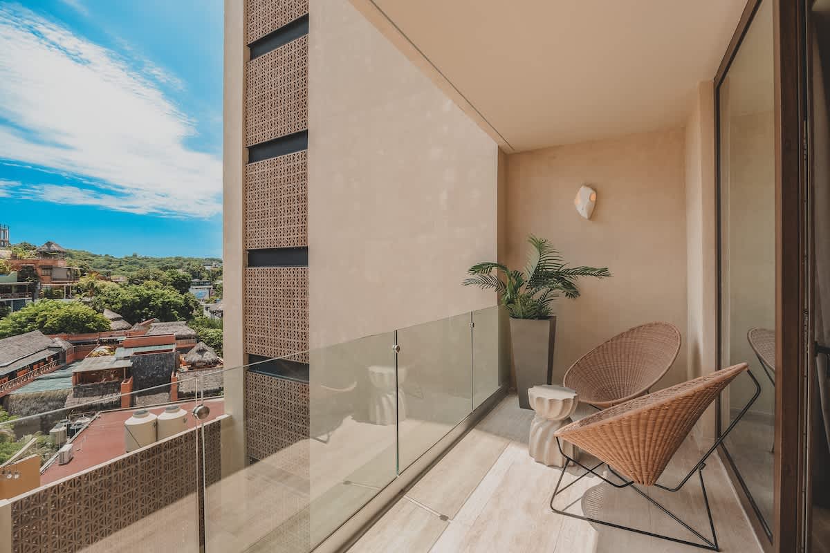 Balcony with two woven chairs, a small side table, and a potted plant, featuring a glass railing and a view of the surrounding buildings and lush tropical foliage.