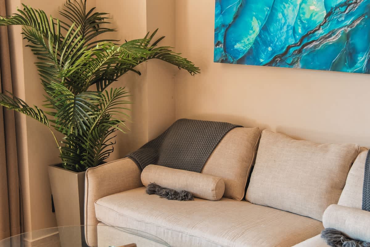 A cozy living room corner features a light beige sofa with a dark gray knitted throw and a bolster pillow, complemented by a tall potted palm plant and a vibrant blue abstract painting on the wall.