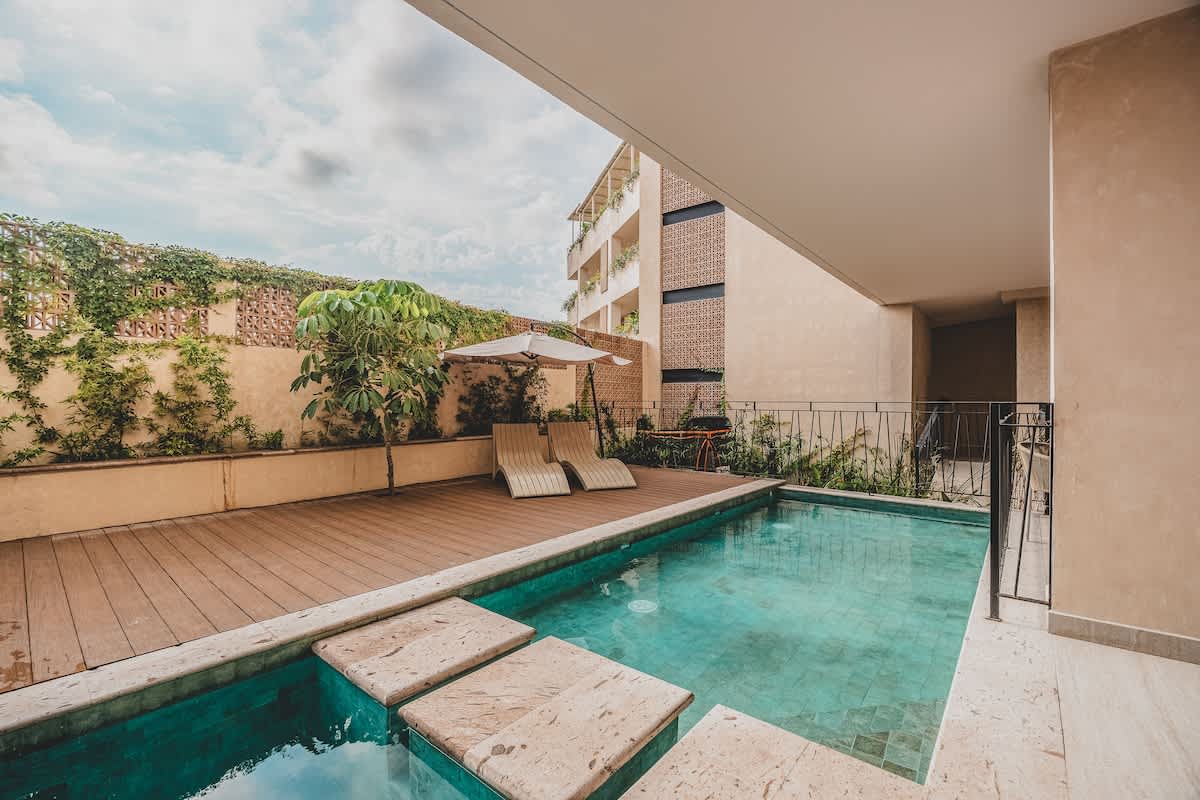 Outdoor swimming pool area featuring a wooden deck with two lounge chairs and a white umbrella, bordered by a stone coping and lush greenery. A modern building with multiple balconies is visible in the background under a partly cloudy sky.