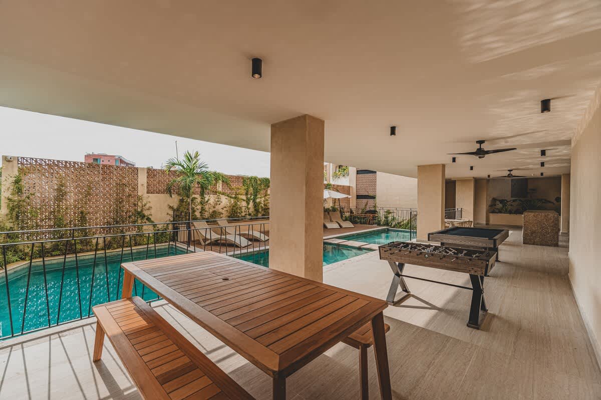 Covered outdoor dining area featuring a long wooden picnic table with matching benches, set on light-colored tiled flooring. A black metal railing offers views of the swimming pool and lush greenery beyond.
