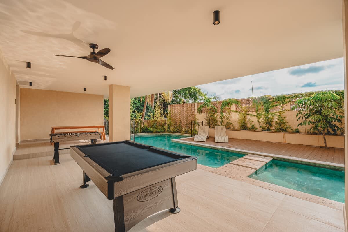 Covered outdoor game area featuring a black-felt pool table and a foosball table on light-colored tiled flooring, with a swimming pool and lush greenery visible beyond. Ceiling fans and recessed lighting are installed overhead.