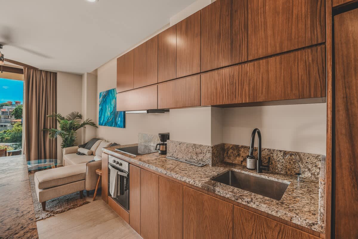 Sleek kitchen with dark wood upper and lower cabinets, granite countertops, and a built-in electric cooktop and oven, seamlessly connecting to the living area with a view of the balcony.