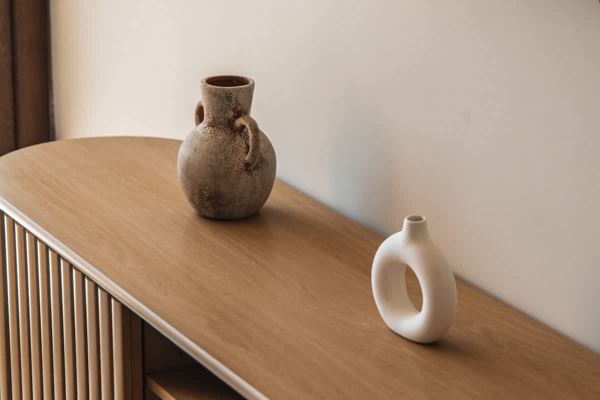 A light wood console table with slatted details holds two decorative vases: a rustic, textured brown vase with handles and a small, smooth white donut-shaped vase.