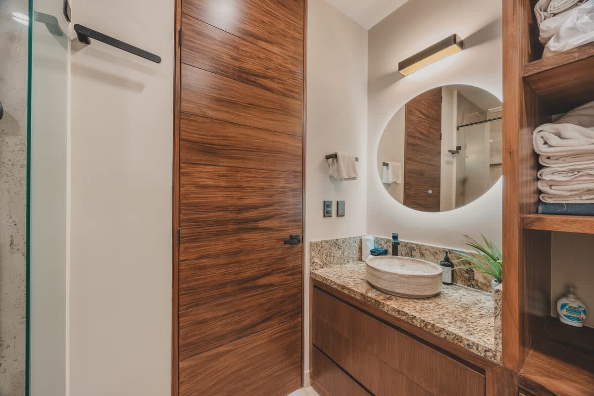 A close-up of the bathroom vanity with a stone vessel sink, granite countertop, and a round backlit mirror, adjacent to a sleek wooden door. Towels are neatly folded on a rack.