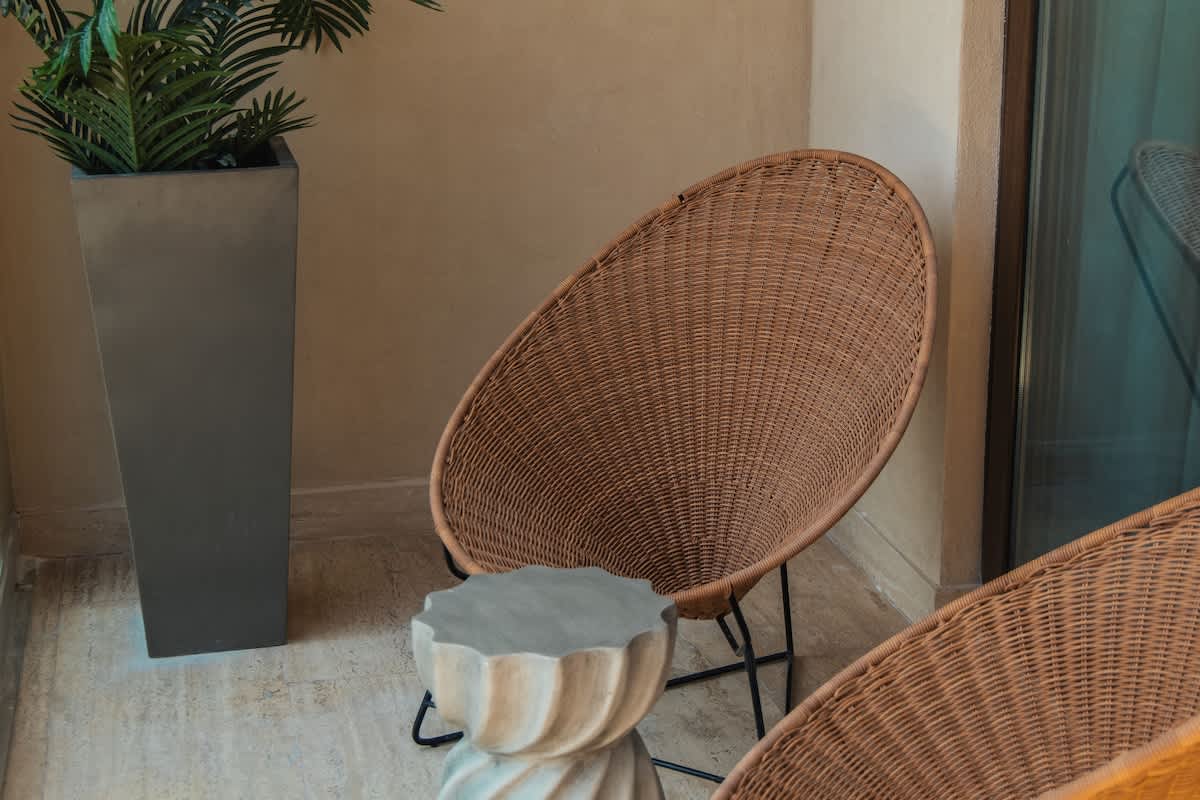 A cozy outdoor seating area on a balcony featuring two round, woven wicker chairs with black metal frames, a unique fluted concrete side table, and a tall planter with a vibrant green plant.