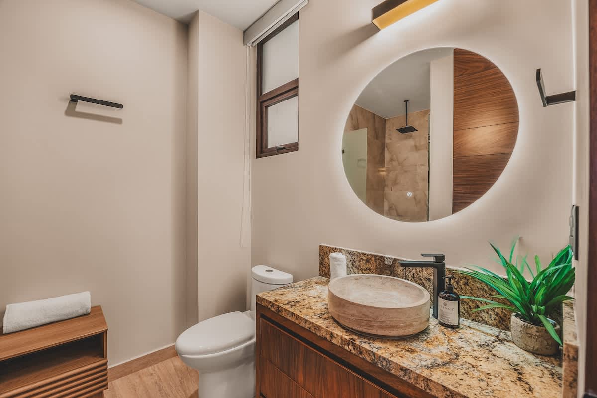 A bright bathroom featuring a toilet next to a vanity with a stone vessel sink, granite countertop, and a round backlit mirror. A small wooden bench or shelf is positioned beneath a window.