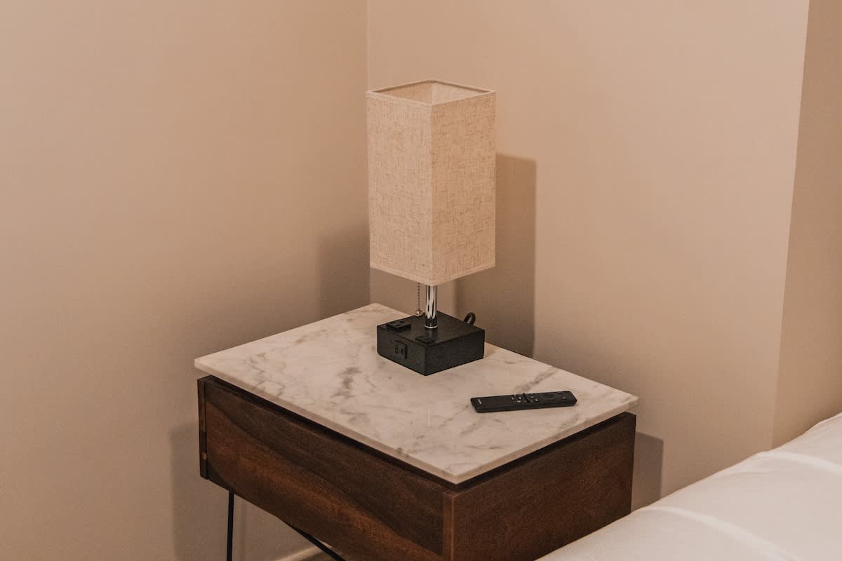 Close-up of a modern bedside table with a white marble top and dark wood drawer, featuring a rectangular fabric lamp and a black remote control, set against a warm beige wall.