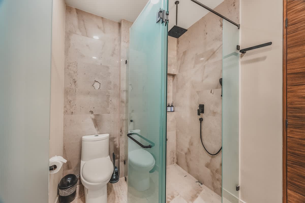 A modern bathroom featuring a toilet enclosed by a frosted glass partition, and a spacious walk-in shower with a rain shower head and handheld sprayer, all set against light marble-look tiles.