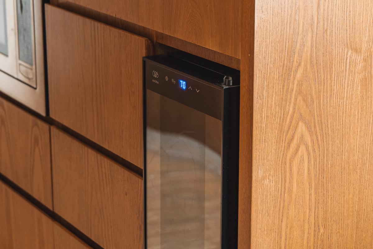 A sleek black beverage cooler with a glass door and digital display is built into light wood cabinetry, adjacent to a microwave, indicating a well-equipped kitchen or bar area.