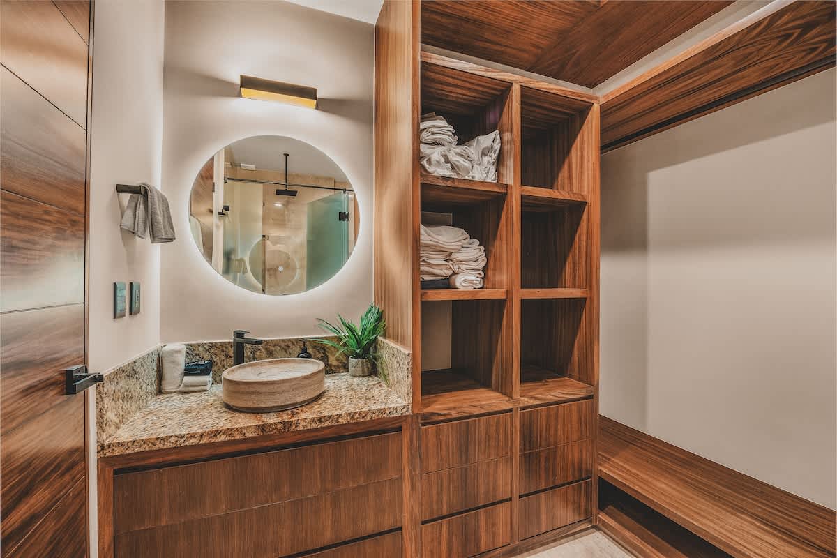 A detailed view of the bathroom vanity area, highlighting the stone vessel sink, granite countertop, and a round backlit mirror, with extensive wooden shelving stocked with neatly folded towels.