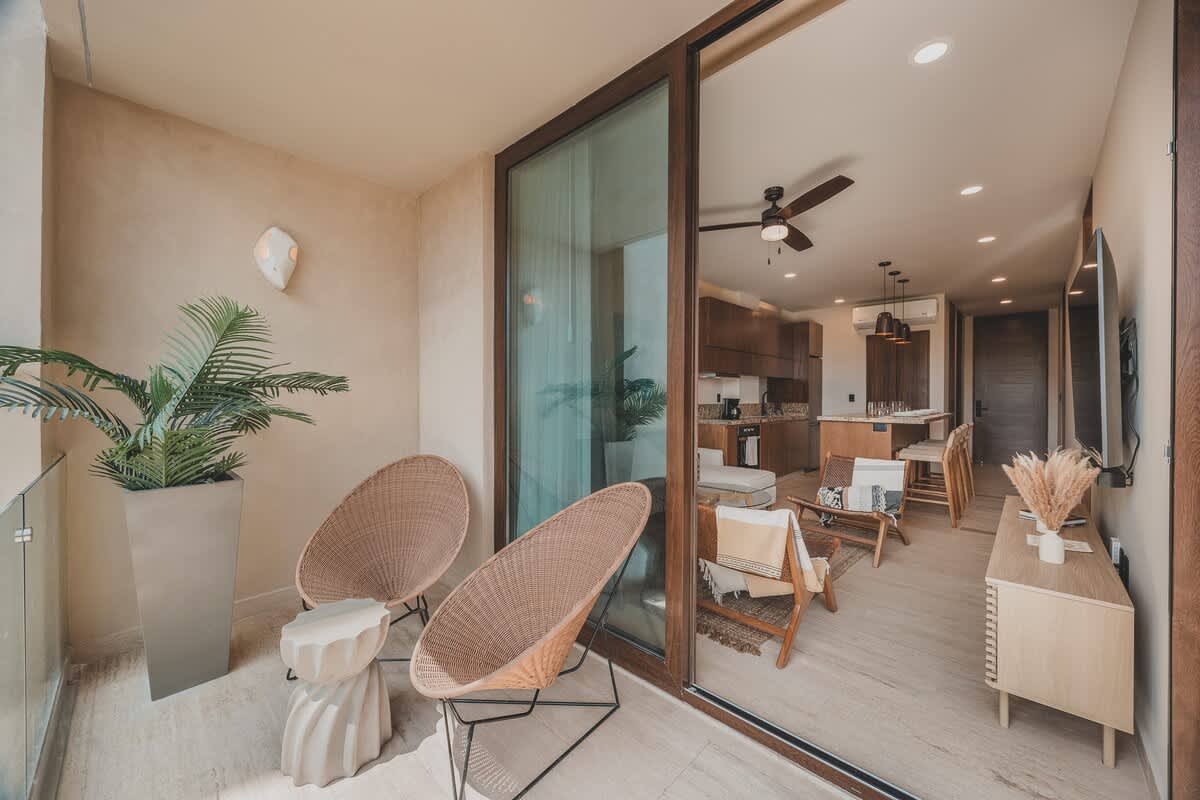 Balcony with two woven chairs and a potted plant, offering a view into an open-concept living area with a kitchen and dining space through large sliding glass doors.