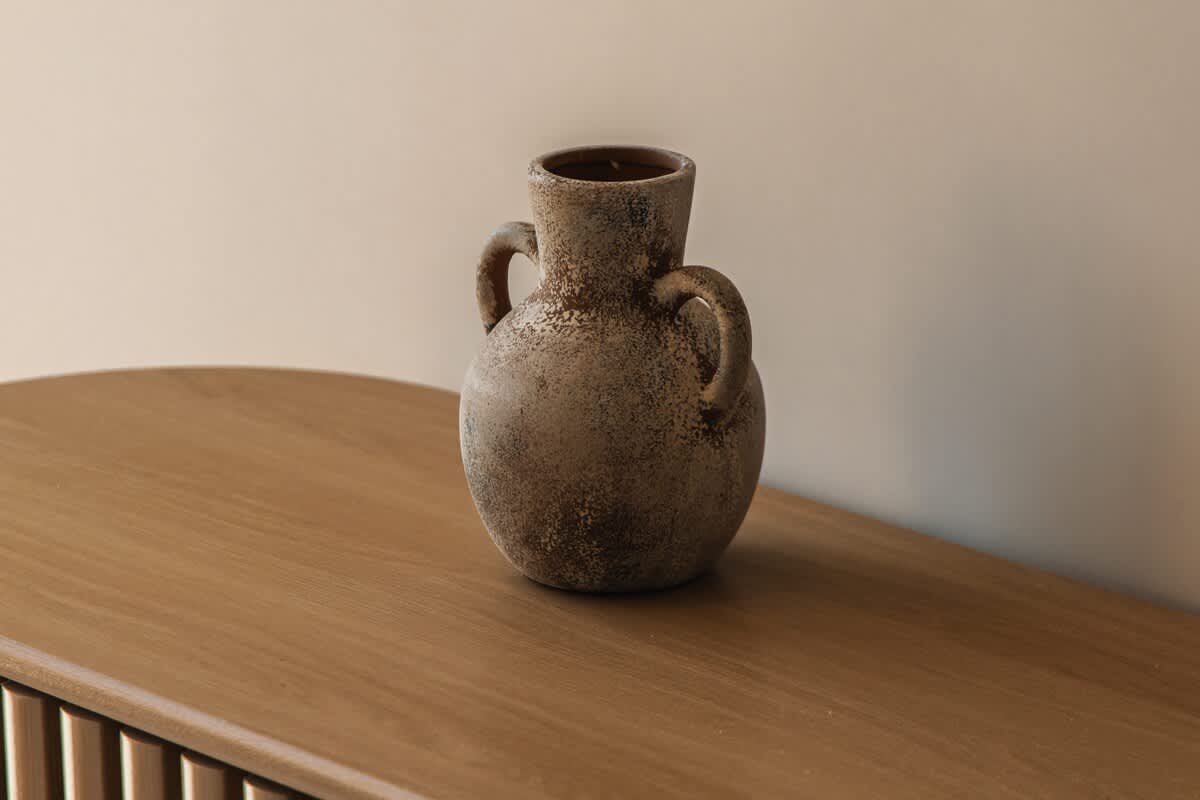 A close-up of a rustic, textured brown ceramic vase with two small handles, sitting on a light wooden console table with vertical slatted details.