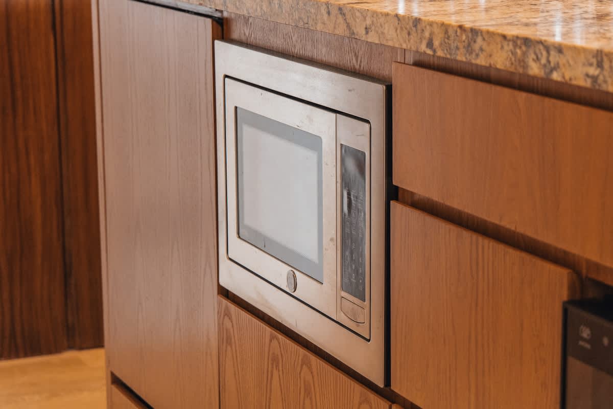 A stainless steel built-in microwave is integrated into light wood cabinetry, set against a speckled granite countertop, suggesting a modern kitchen or kitchenette design.