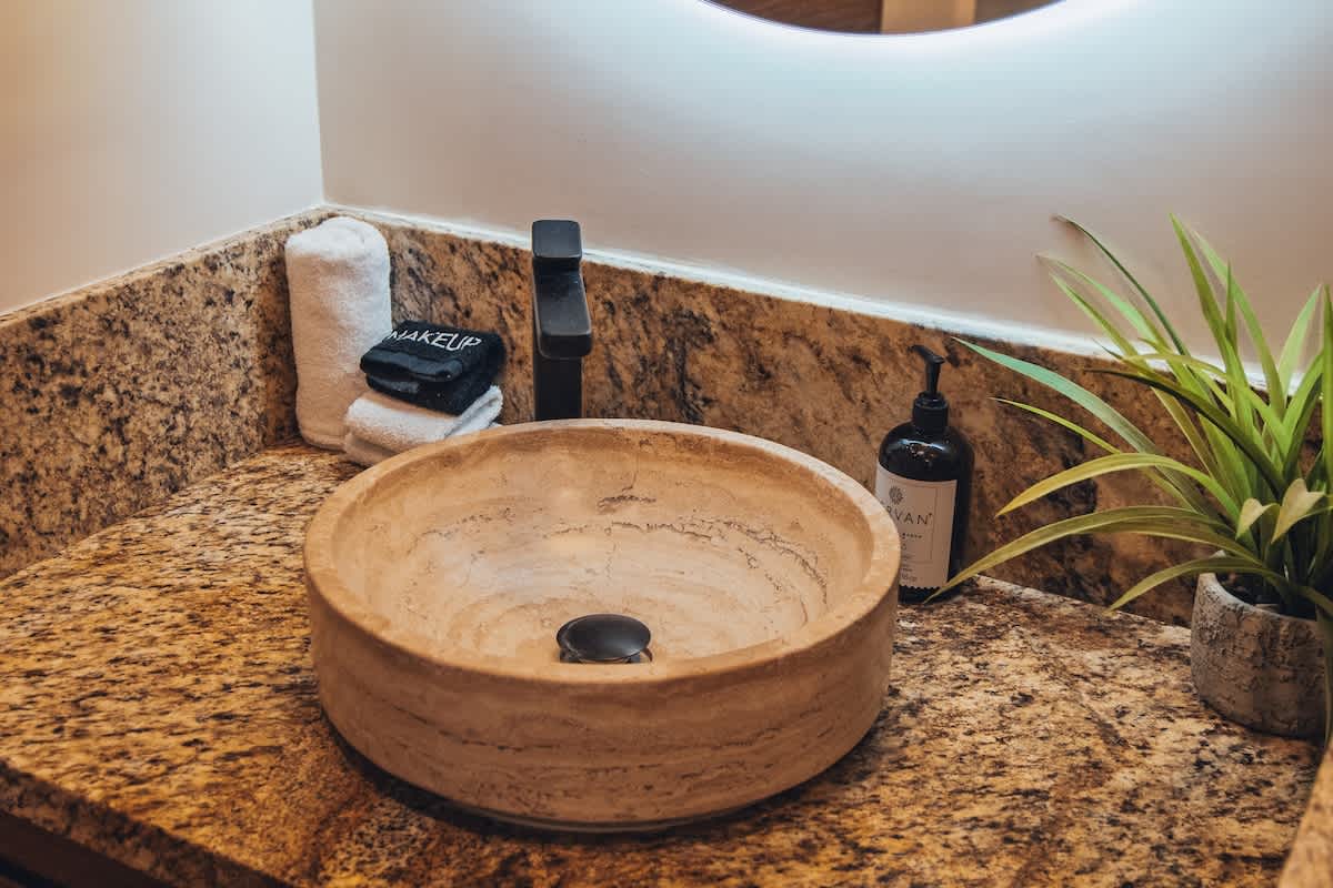A round, light-colored vessel sink with a black faucet is centered on a speckled granite countertop, flanked by a black soap dispenser, rolled white towels, and a small plant.