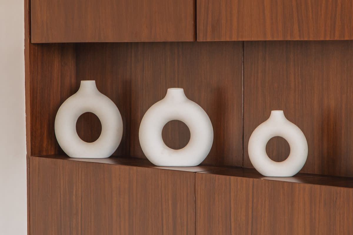 Three minimalist white ceramic vases, each with a circular opening in the center, are displayed on a dark wooden shelf.