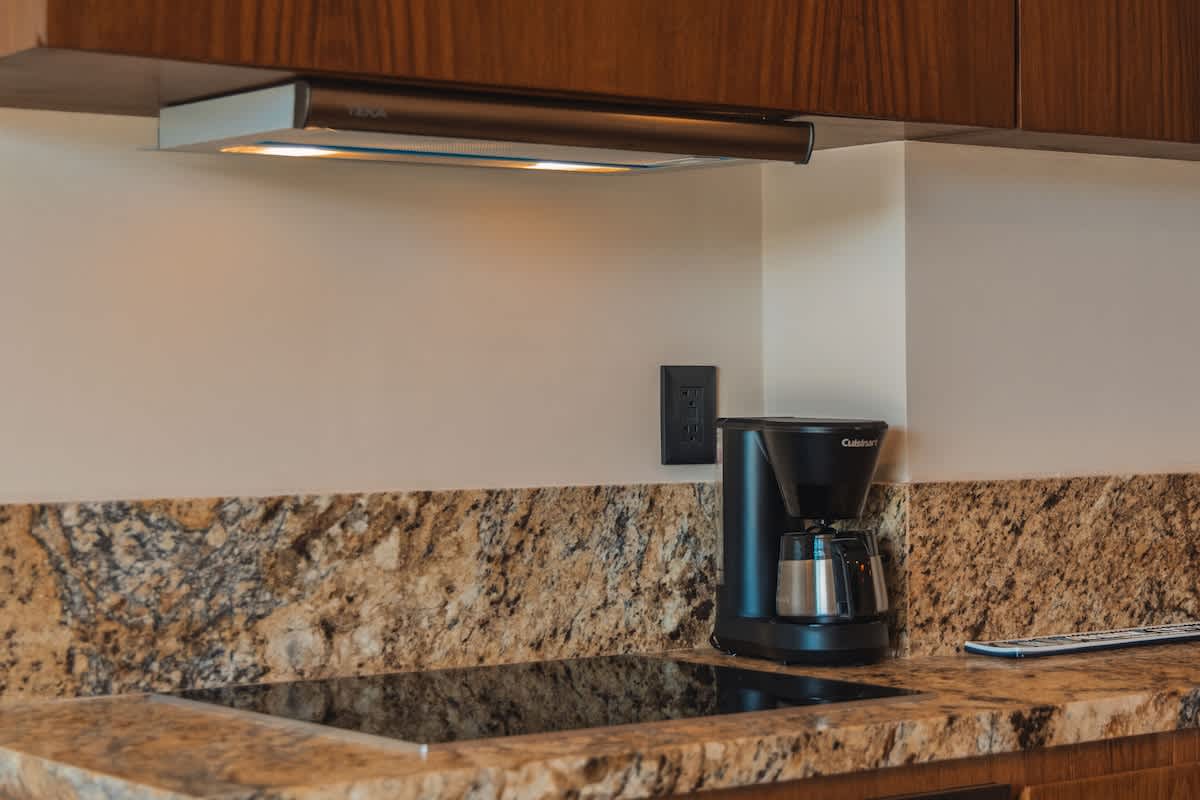 A black coffee maker sits on a speckled granite countertop next to a black induction cooktop, with a stainless steel range hood and wooden cabinetry visible above.