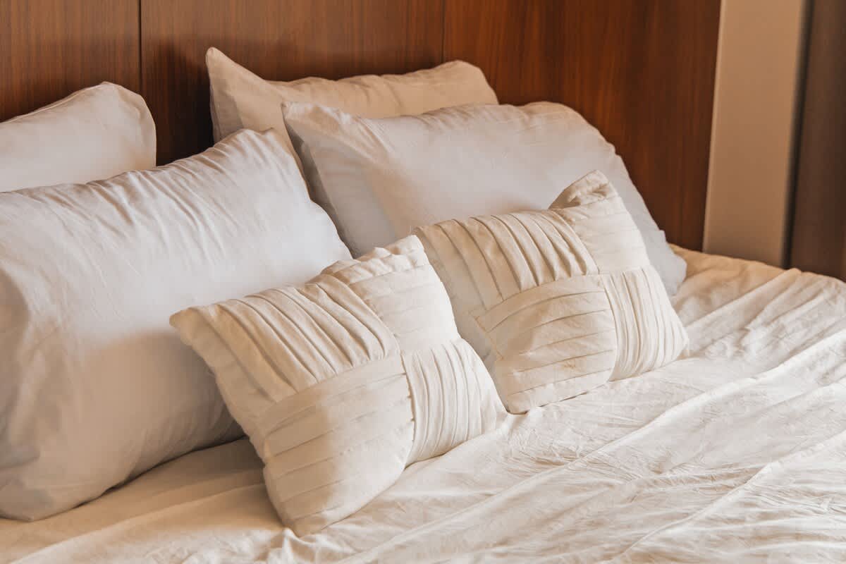 A close-up of a bed with multiple white pillows, including two decorative pillows with pleated textures, set against a dark wood headboard.