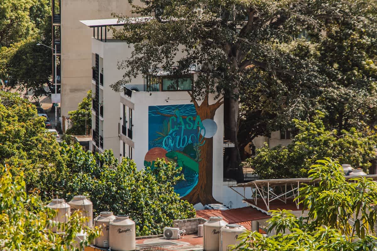 The exterior of a multi-story building is adorned with a vibrant, large-scale mural depicting a stylized tree and an ocean scene, blending art with architecture. The building is surrounded by lush green foliage and other urban structures.