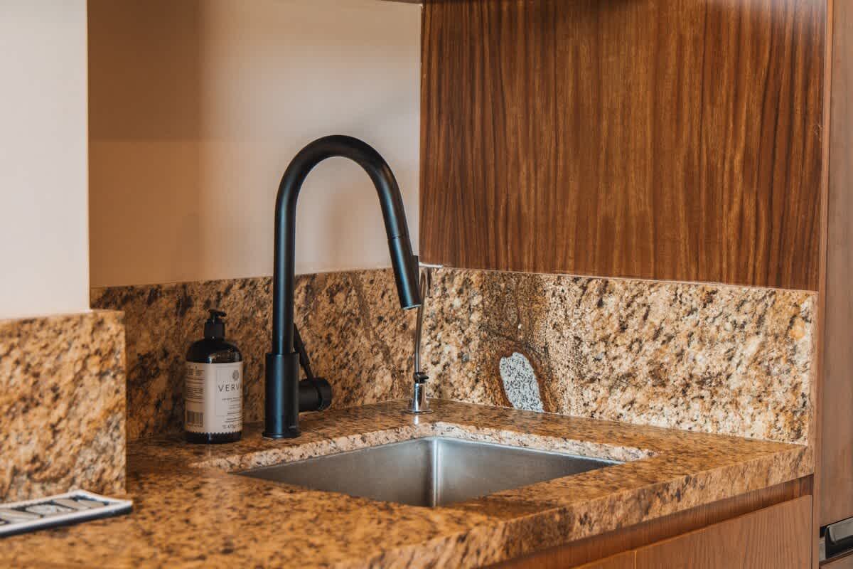 A sleek black gooseneck faucet extends over a stainless steel sink, set into a speckled granite countertop, with a black soap dispenser and rich wooden paneling on the wall.