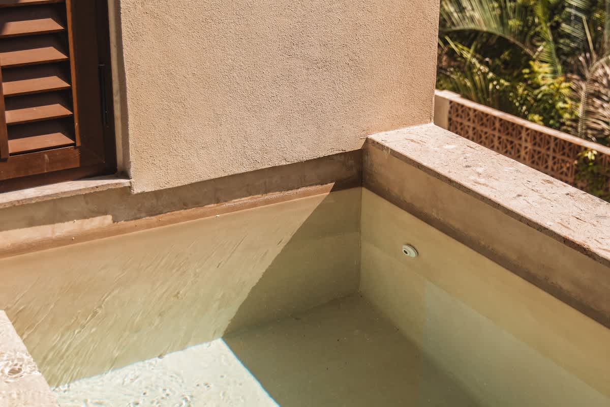 A detailed shot of the corner of a hot tub, showing the light-colored steps leading into the water and a small jet, with a textured wall and wooden shutters in the background.