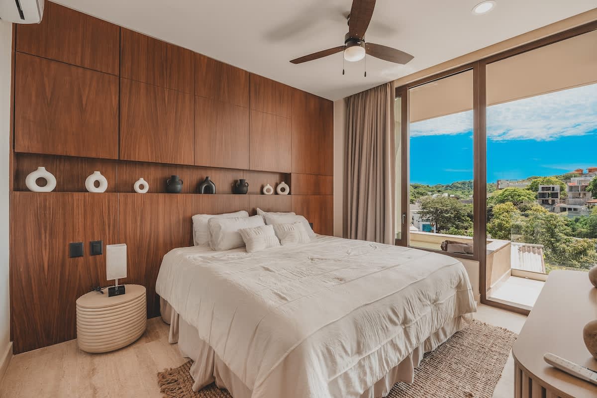 A modern bedroom showcasing a large wooden accent wall with integrated decorative niches above a white-linened bed, flanked by a round nightstand and lamp. Large glass doors offer a scenic view of the outdoors.