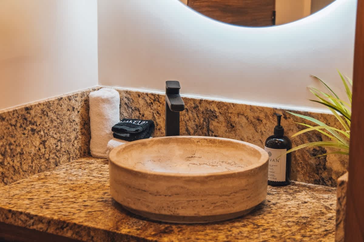 A light-colored vessel sink and modern black faucet are set on a granite countertop, with a black soap dispenser, white towels, and a round illuminated mirror visible above.
