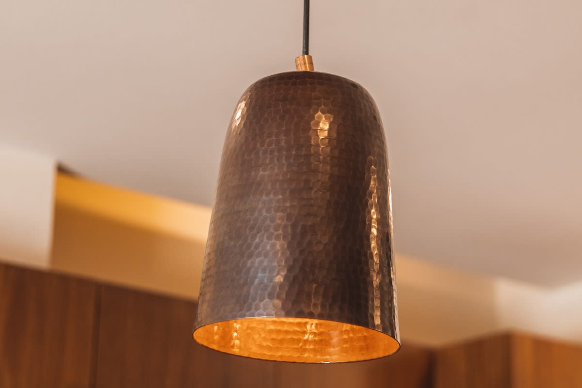 A close-up of a hammered copper pendant light fixture with a warm, reflective interior, hanging from a black cord against a light ceiling.