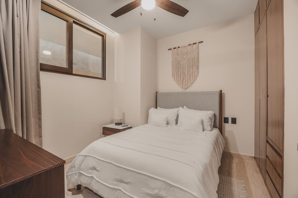 Bedroom with a queen-sized bed, a macrame wall hanging above the headboard, a dark wood dresser, and a built-in wardrobe, illuminated by natural light from a high window.