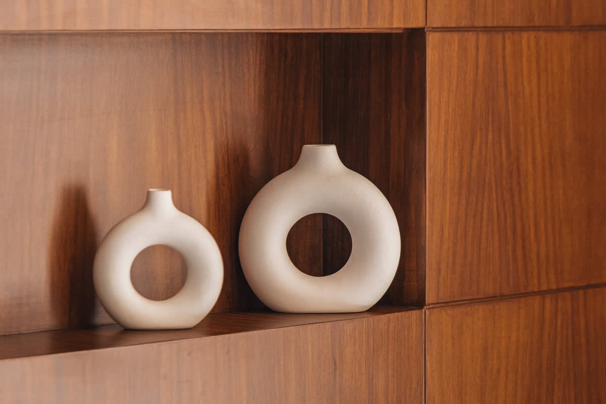 Two minimalist white ceramic vases, one larger and one smaller, both featuring a circular opening, are arranged on a dark wooden shelf.