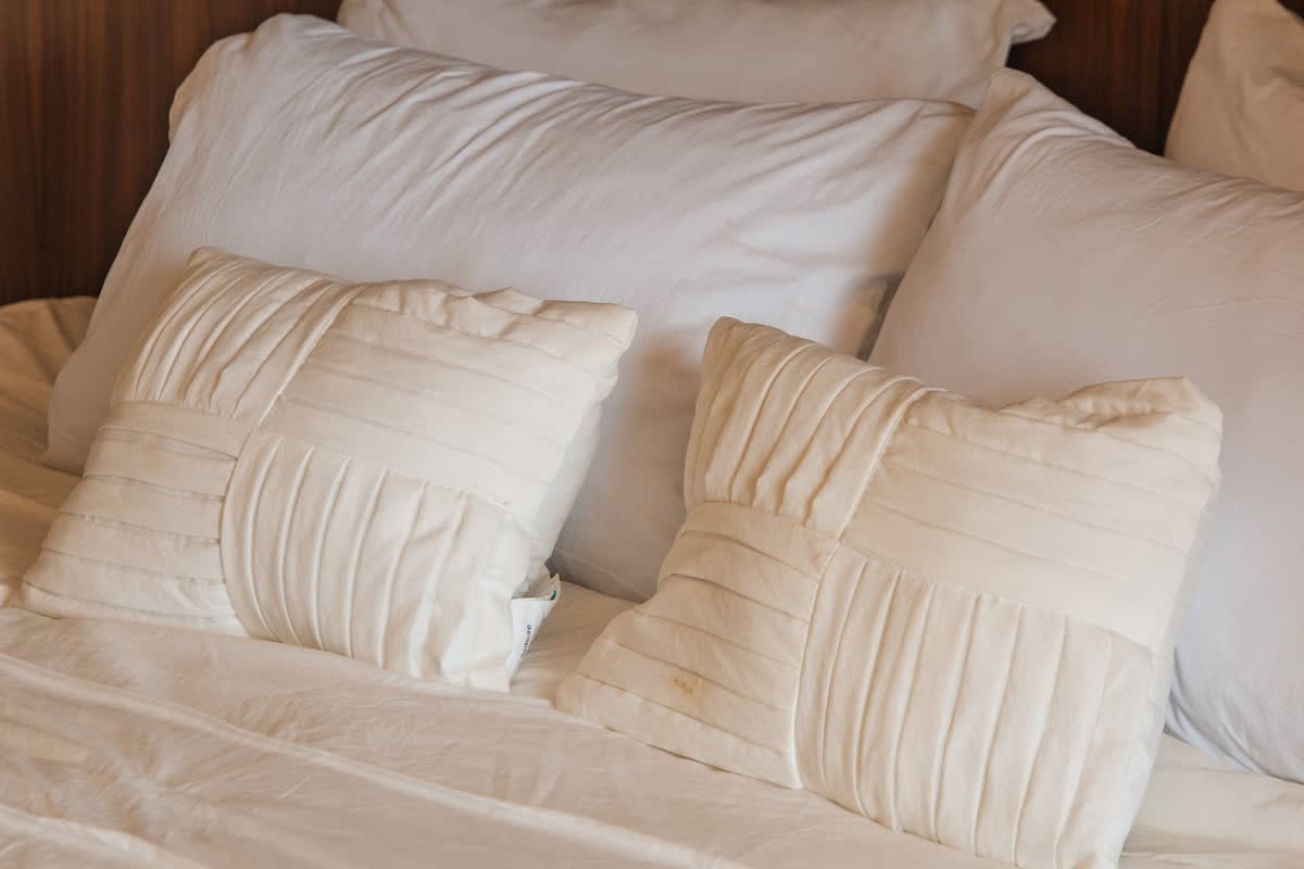 A close-up view of two decorative square pillows with intricate pleated and paneled textures, resting on a bed with crisp white sheets and larger white pillows, enhancing the bed's comfort and style.