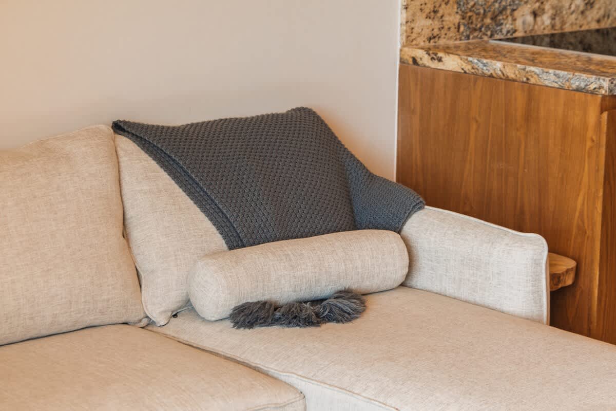 Close-up of a light beige sofa corner with a dark gray knitted throw draped over the back and a matching bolster pillow, set against a light-colored wall and a glimpse of a granite countertop.