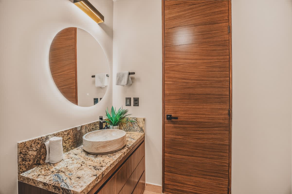 Modern bathroom featuring a circular backlit mirror above a vessel sink set on a granite countertop, with dark wood cabinetry and a matching dark wood door.
