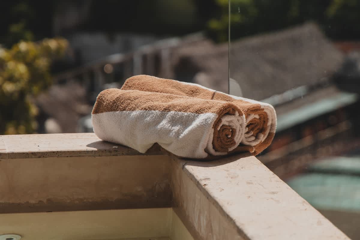 Two neatly rolled brown and white striped towels rest on a light-colored concrete ledge, with a blurred outdoor background suggesting a pool area or balcony with greenery.