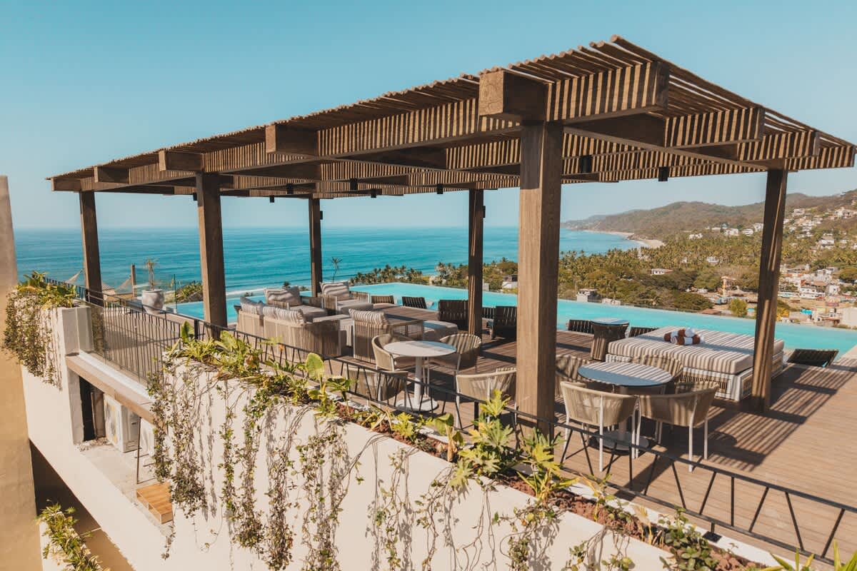 A luxurious rooftop terrace features a large, shaded seating area under a wooden pergola, offering comfortable outdoor furniture. An infinity pool stretches along the edge, providing stunning panoramic views of the ocean and the vibrant coastal town below.