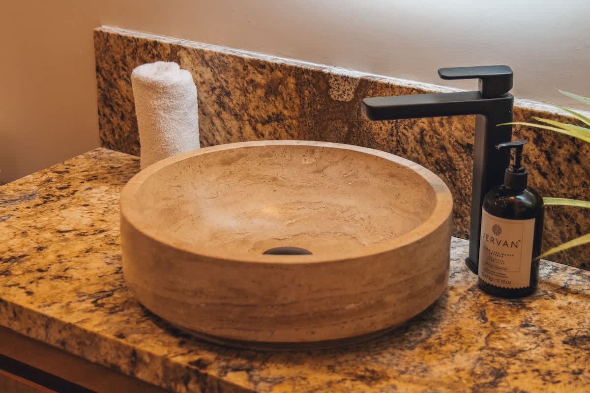 A stylish bathroom vanity features a unique round, light-colored stone vessel sink paired with a modern matte black faucet, set against a speckled granite countertop. A rolled white towel and a black soap dispenser complete the elegant setup.