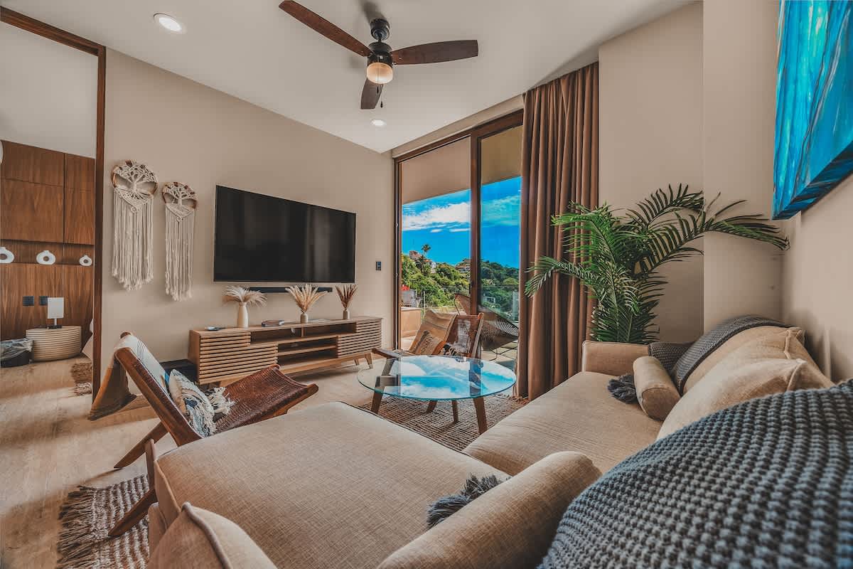 Cozy living room with a large L-shaped sectional sofa, a glass coffee table, and two woven lounge chairs, facing a wall-mounted TV and a sliding glass door leading to a balcony with lush views.