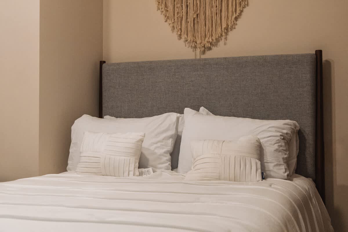 A neatly made bed with a grey upholstered headboard, crisp white sheets, and multiple white pillows, including two decorative square pillows with textured patterns, beneath a macrame wall hanging.