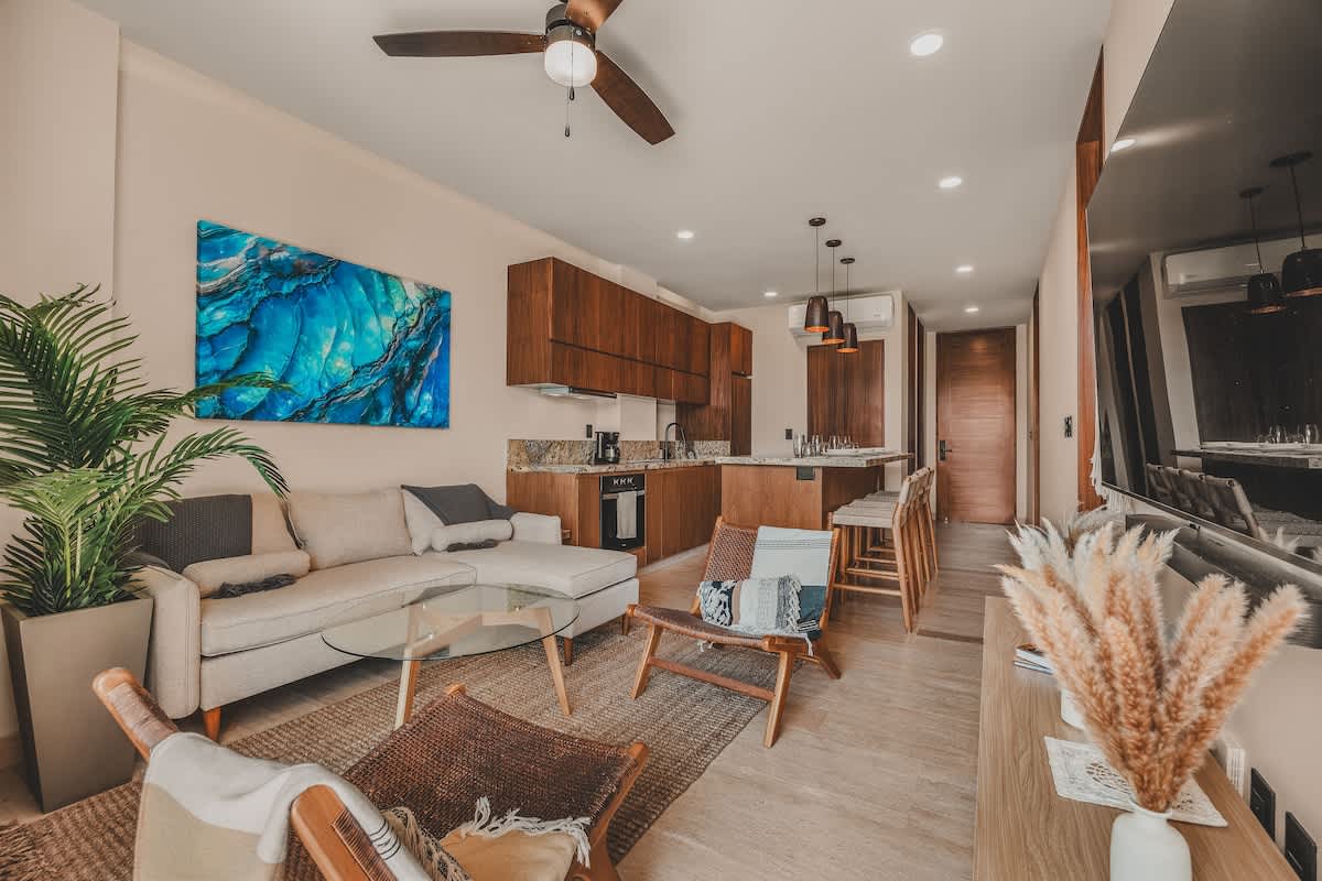 Expansive open-concept living and kitchen area, featuring a light-colored sectional sofa, a glass coffee table, and a wall-mounted TV, with a full kitchen island and dark wood cabinetry visible in the background.