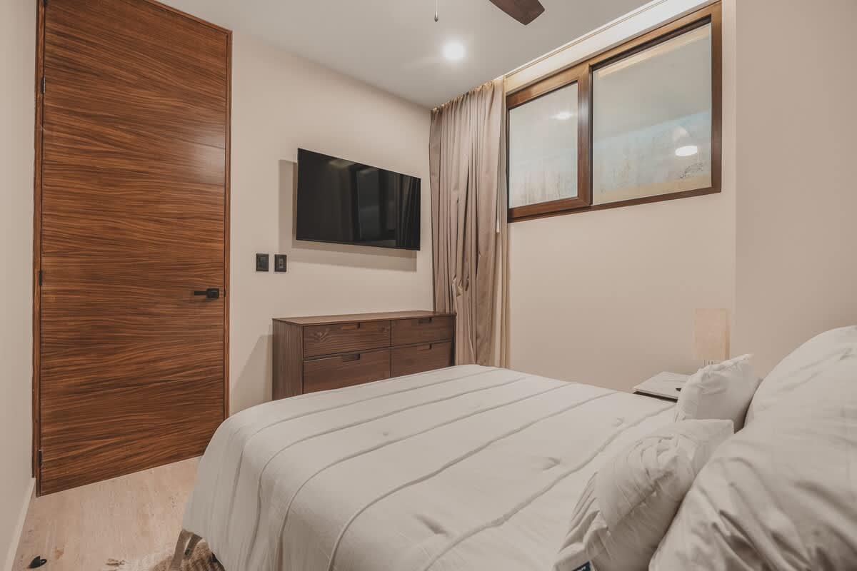 A cozy bedroom featuring a wall-mounted TV above a dark wooden dresser, a bed with white linens, and a striking wooden door with horizontal grain. A small window with a wooden frame is visible on the right.