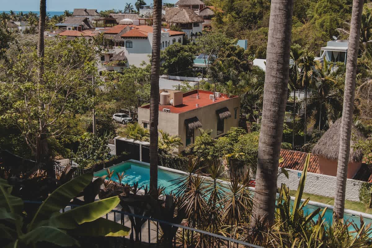 Elevated perspective from a balcony, offering a panoramic view of a swimming pool, various residential buildings with traditional red tile roofs, and abundant tropical trees, with the ocean visible on the horizon.