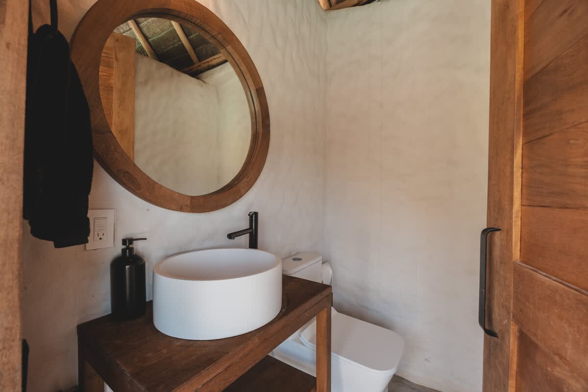 A compact bathroom features a white vessel sink on a rustic wooden vanity, a modern toilet, and a large round wooden-framed mirror, with white textured walls and a thatched ceiling visible.